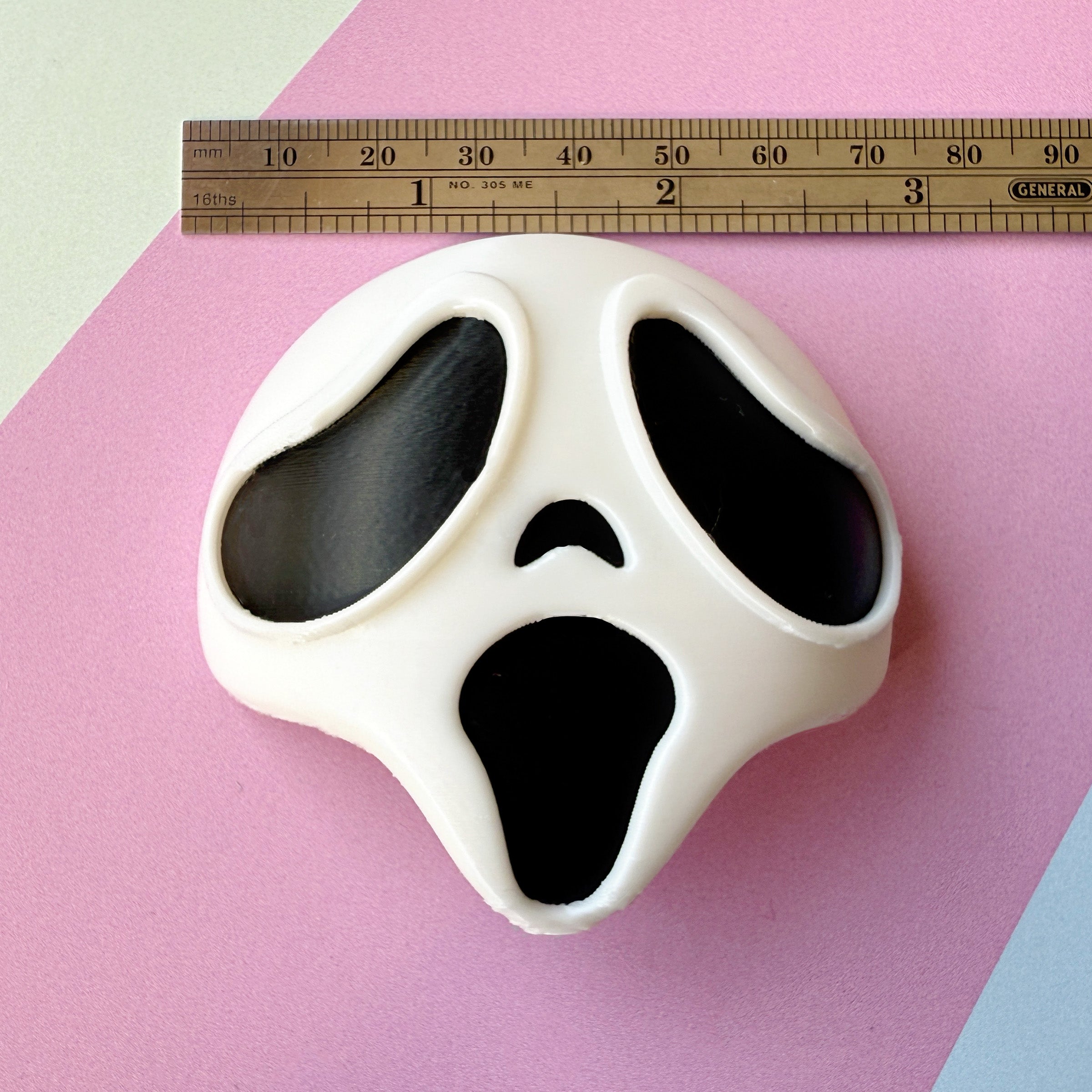 White mask with black eyes and mouth design on a pink background, ruler for scale.