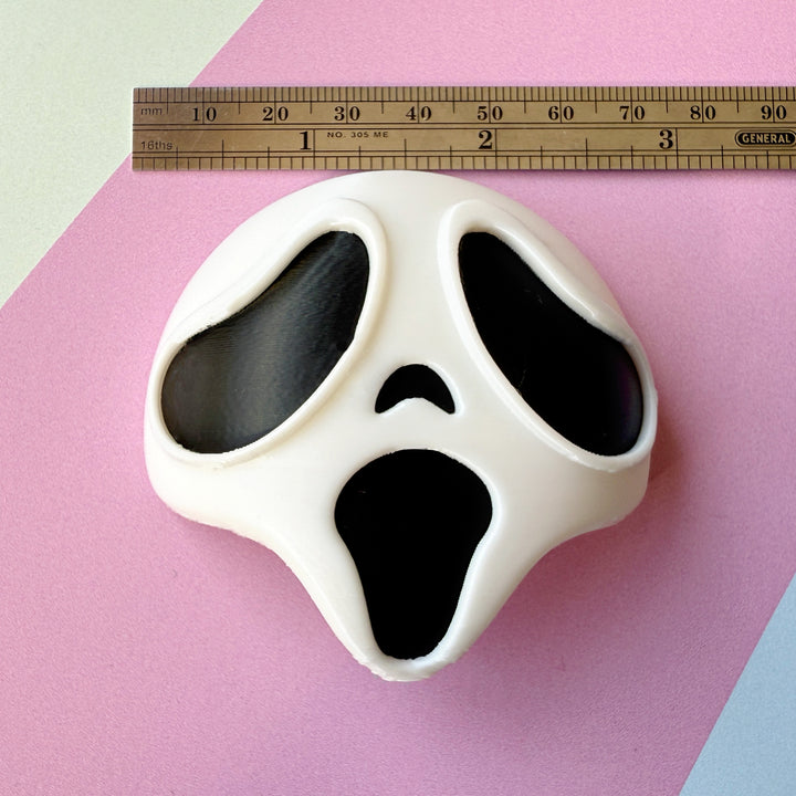 White mask with black eyes and mouth design on a pink background, ruler for scale.