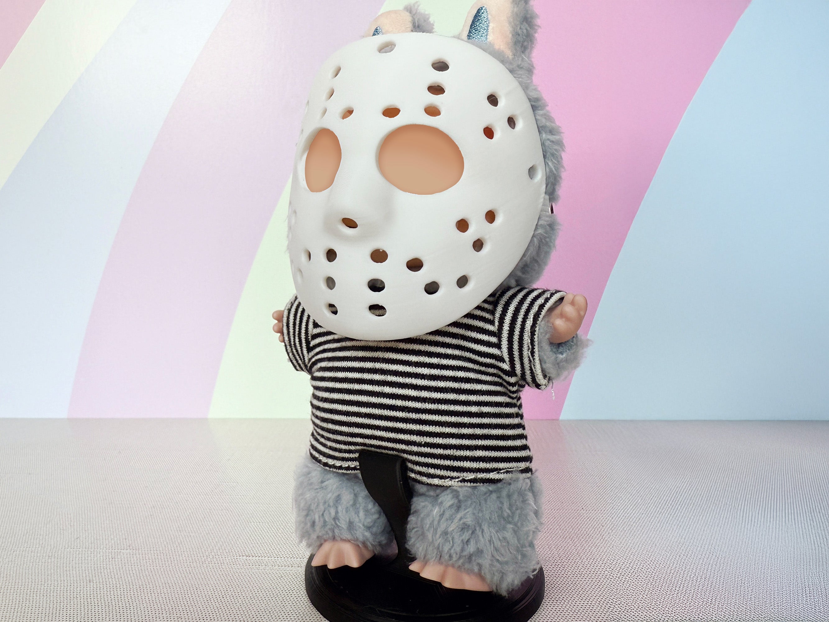 Plush toy with a Miniature hockey-style mask, 3D printed by Necessary Design., wearing a striped shirt against a colorful background.