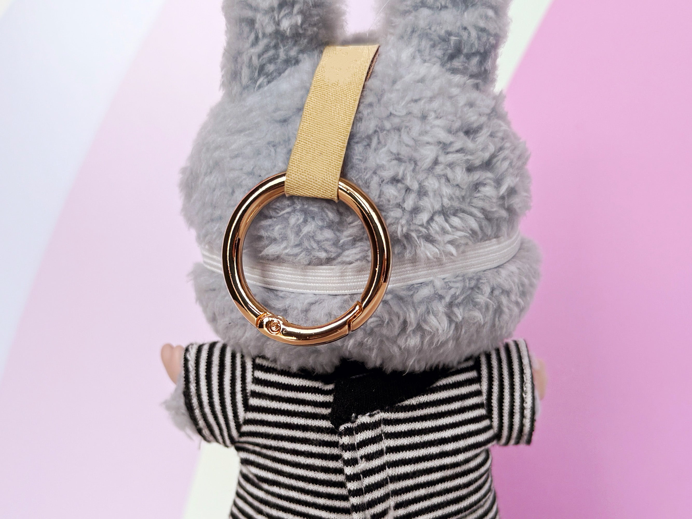 Back of Plush toy wearing hockey-style mask secured with white elastic strap with a keychain ring and beige strap on a pink background.