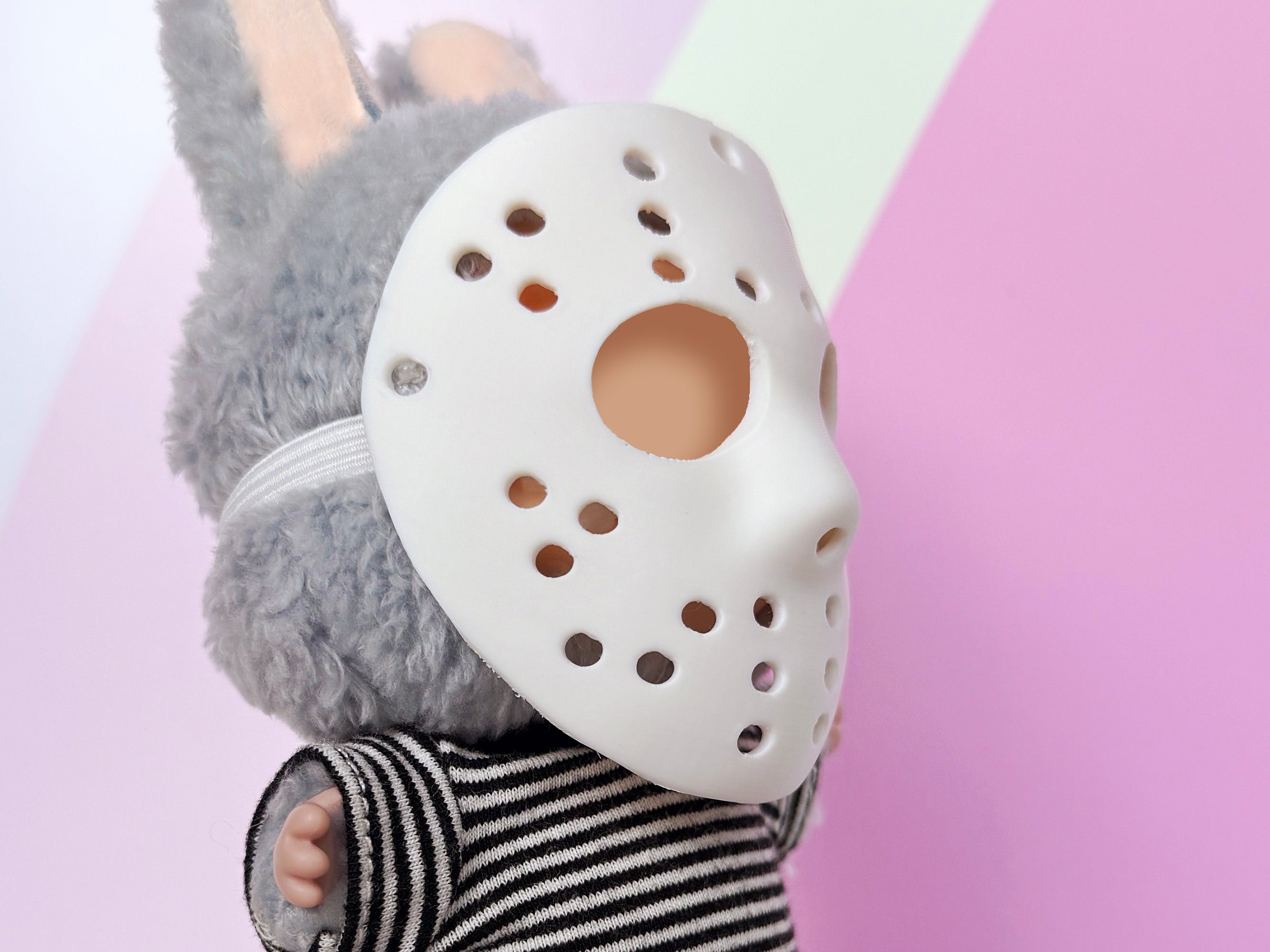 Side view of Plush toy with a Miniature hockey-style mask, 3D printed by Necessary Design., wearing a striped shirt against a colorful background.