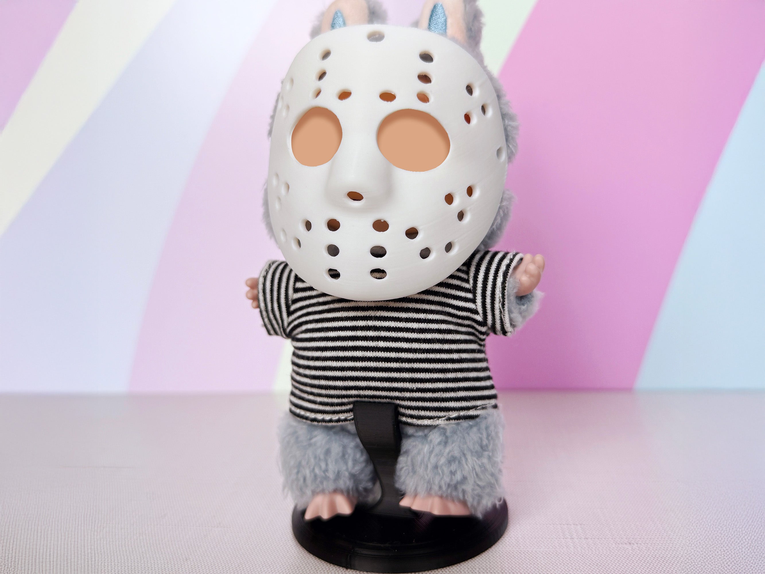 Front view of Plush toy with a Miniature hockey-style mask, 3D printed by Necessary Design., wearing a striped shirt on a black display stand against a colorful background.