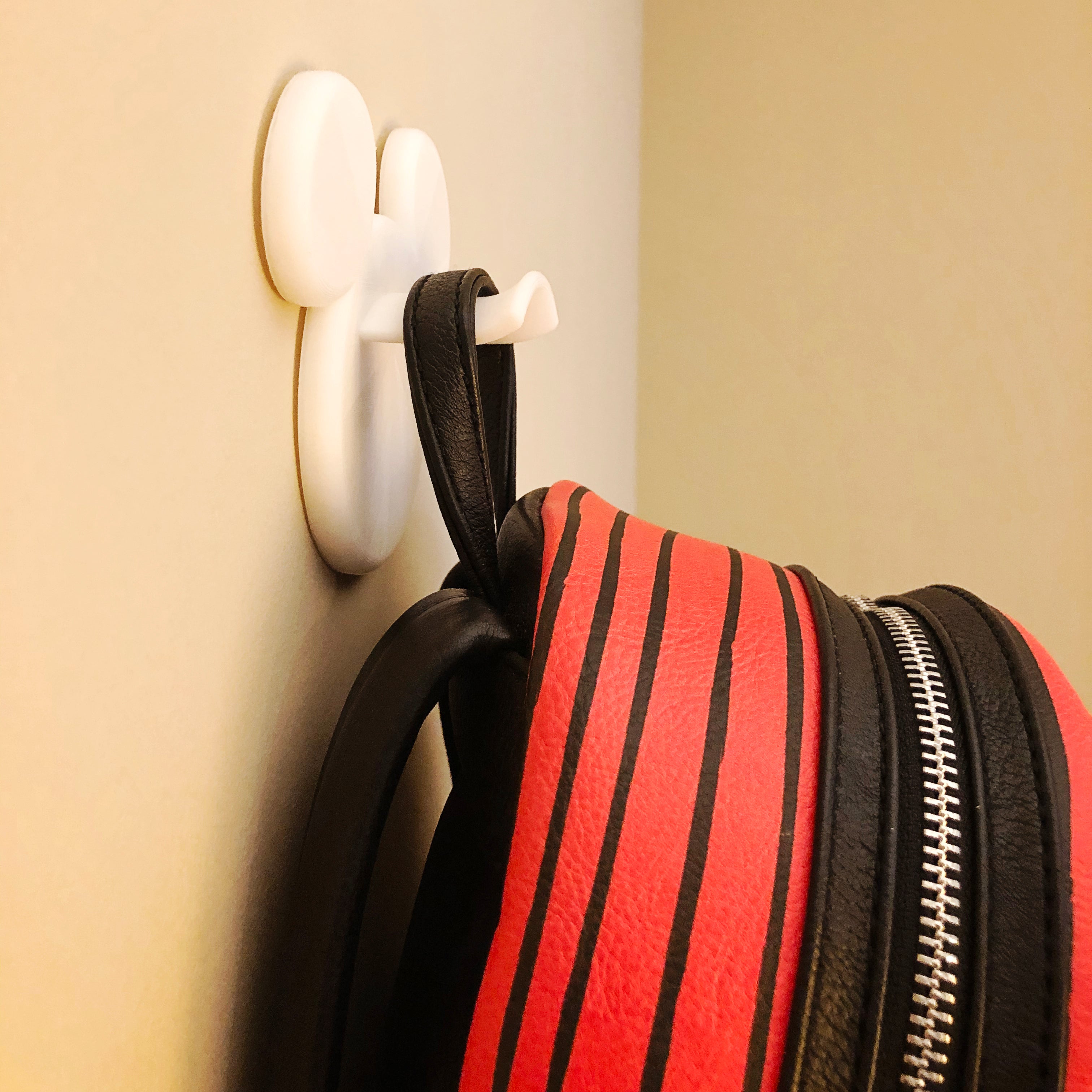 White wall hook holding a red and black striped bag against a beige wall.