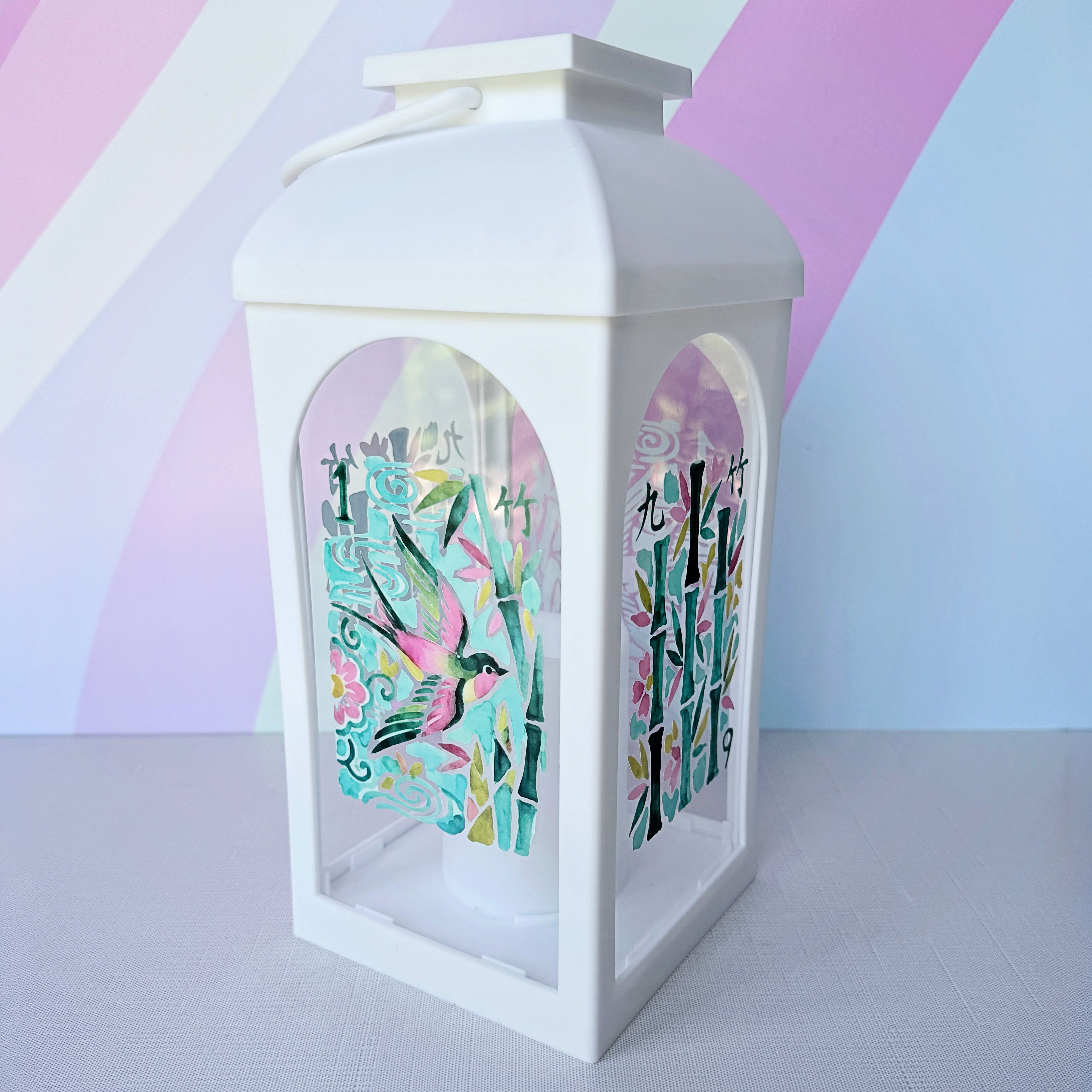 A white decorative lantern with arched side panels, featuring colorful printed Japanese-style artwork with birds, bamboo, and characters. The lantern is photographed on a light surface against a pastel striped background.