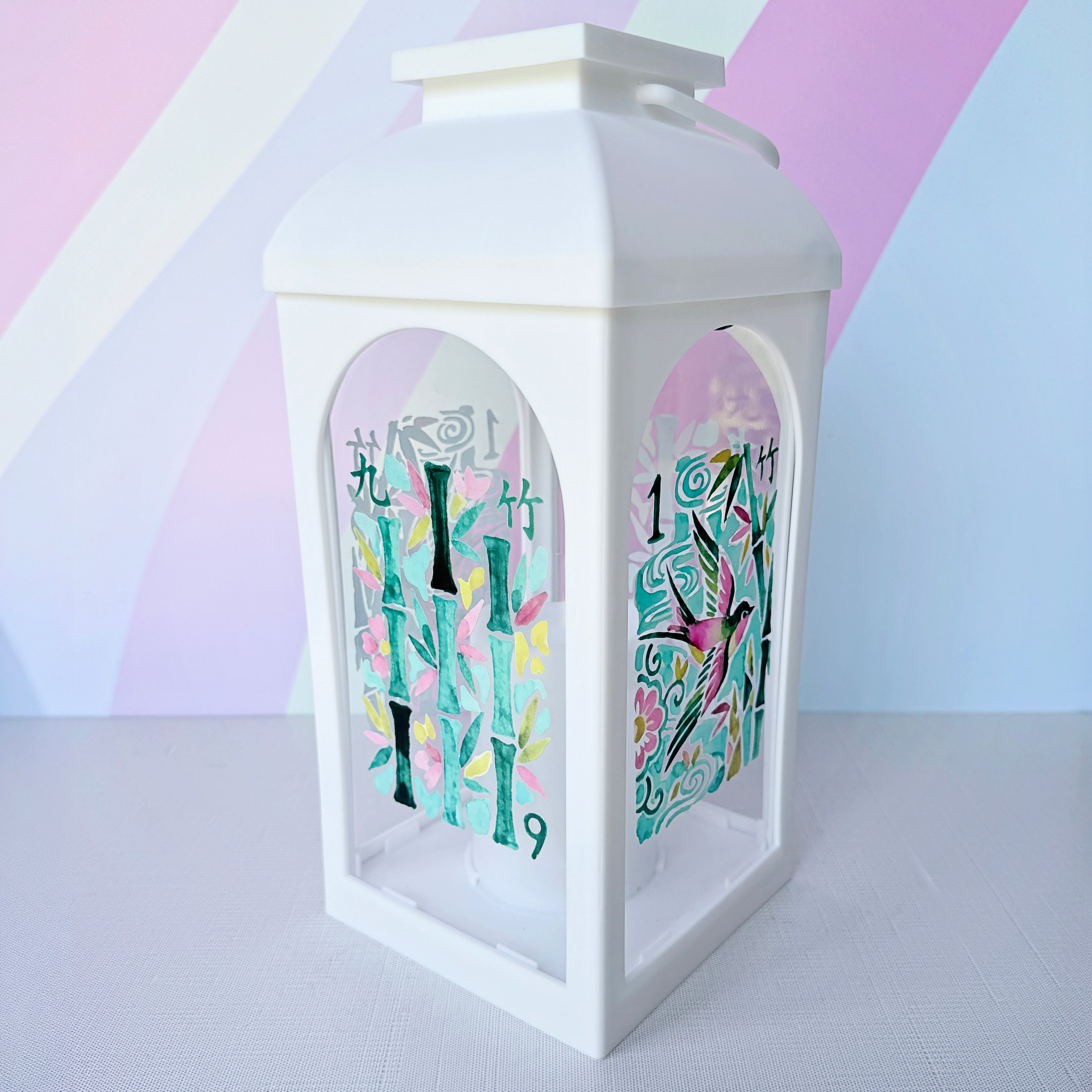A white lantern with arched side windows, featuring colorful printed bamboo and bird illustrations, set against a pastel striped background.