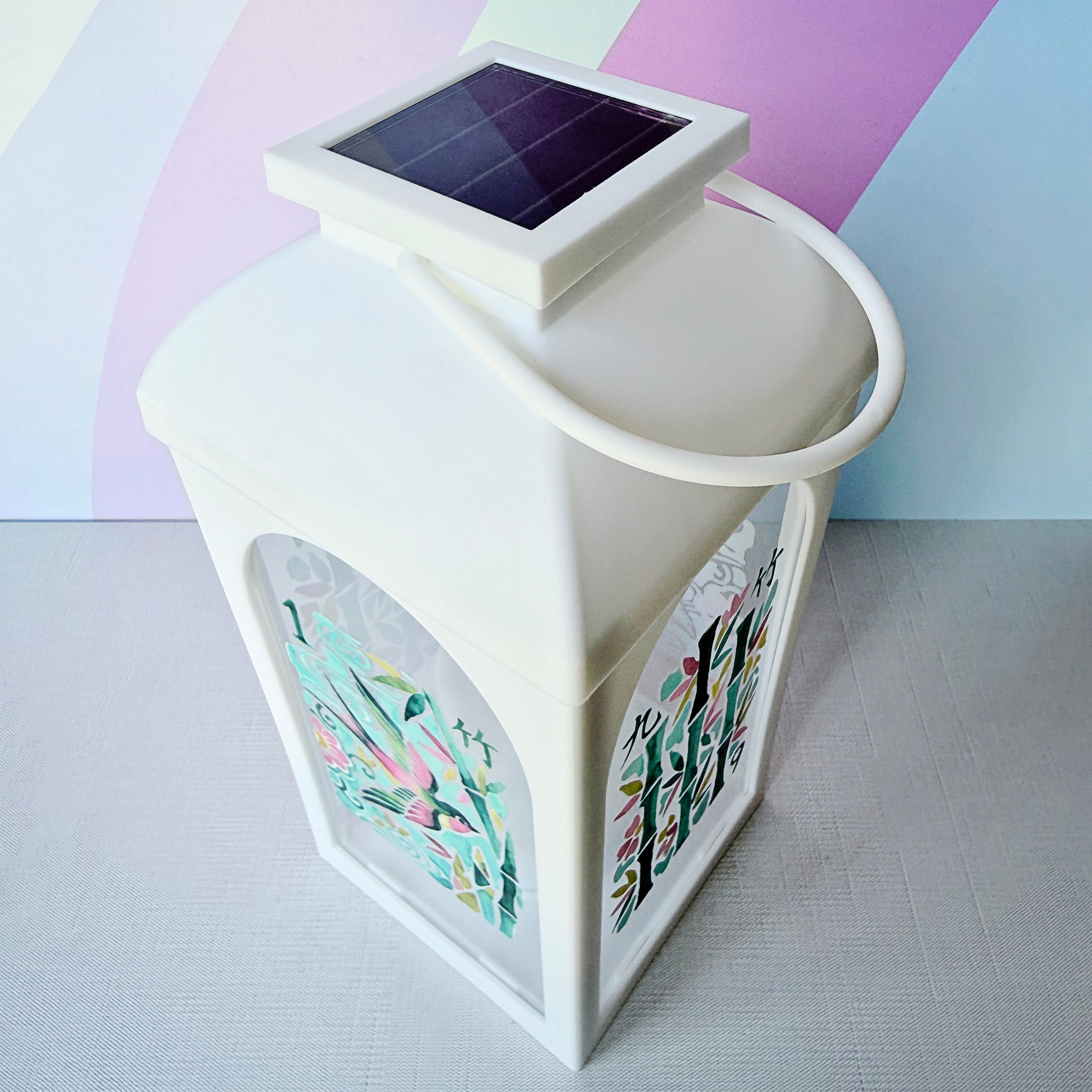 A white solar-powered lantern with a handle and a small solar panel on top, featuring colorful printed floral designs on its sides, set against a pastel rainbow background.