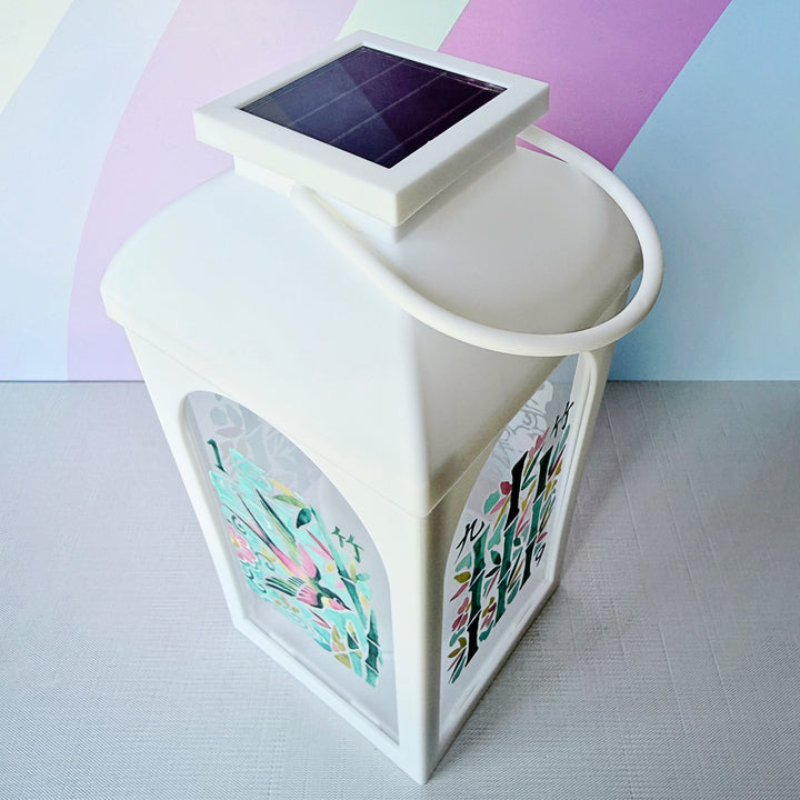 A white solar-powered lantern with a handle and a small solar panel on top, featuring colorful printed floral designs on its sides, set against a pastel rainbow background.