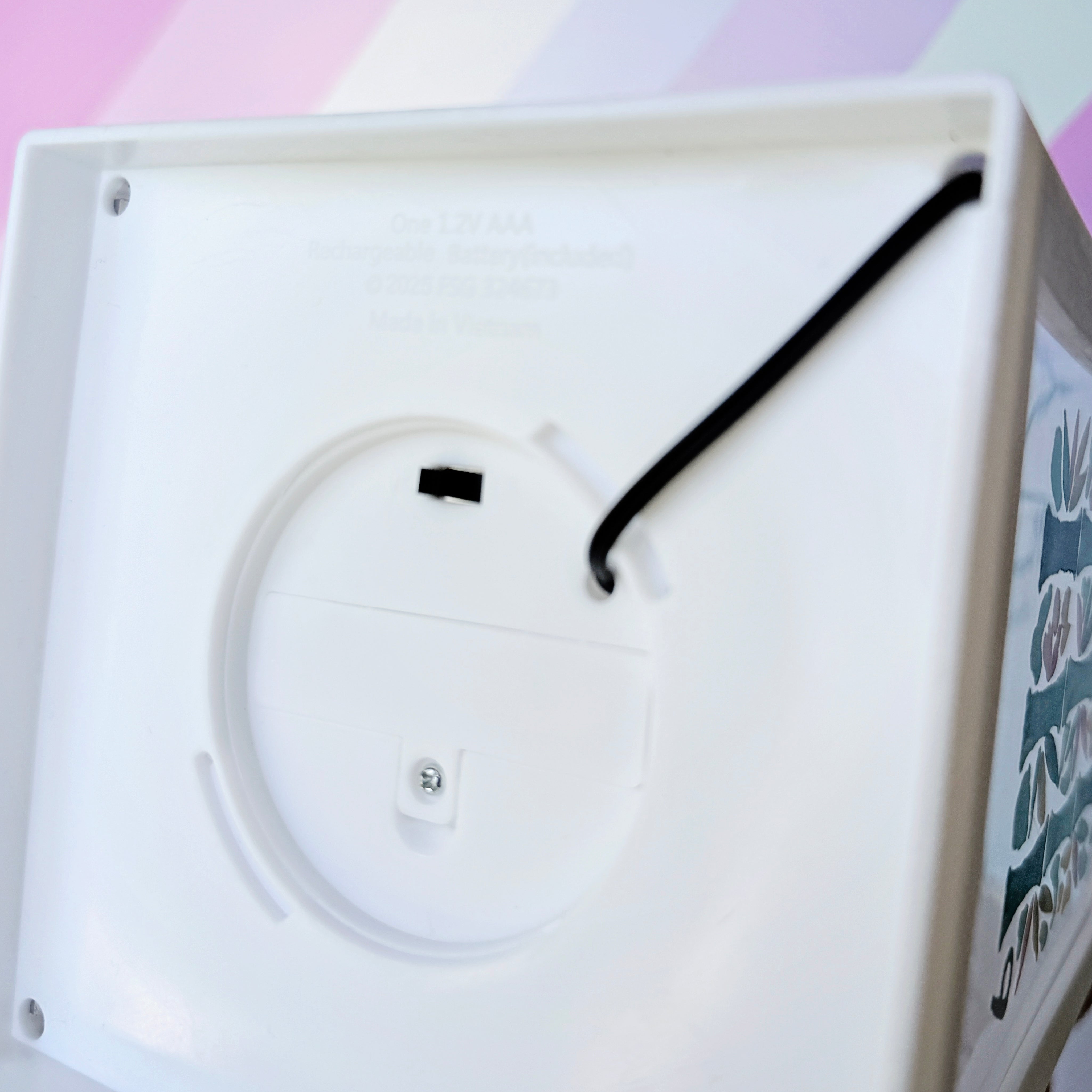 Close-up of the underside of a white plastic lantern, showing a circular recessed area, a small black component, and a black cord looped through a hole. Faint text is visible inside the plastic, and part of a colorful side panel appears on the right.