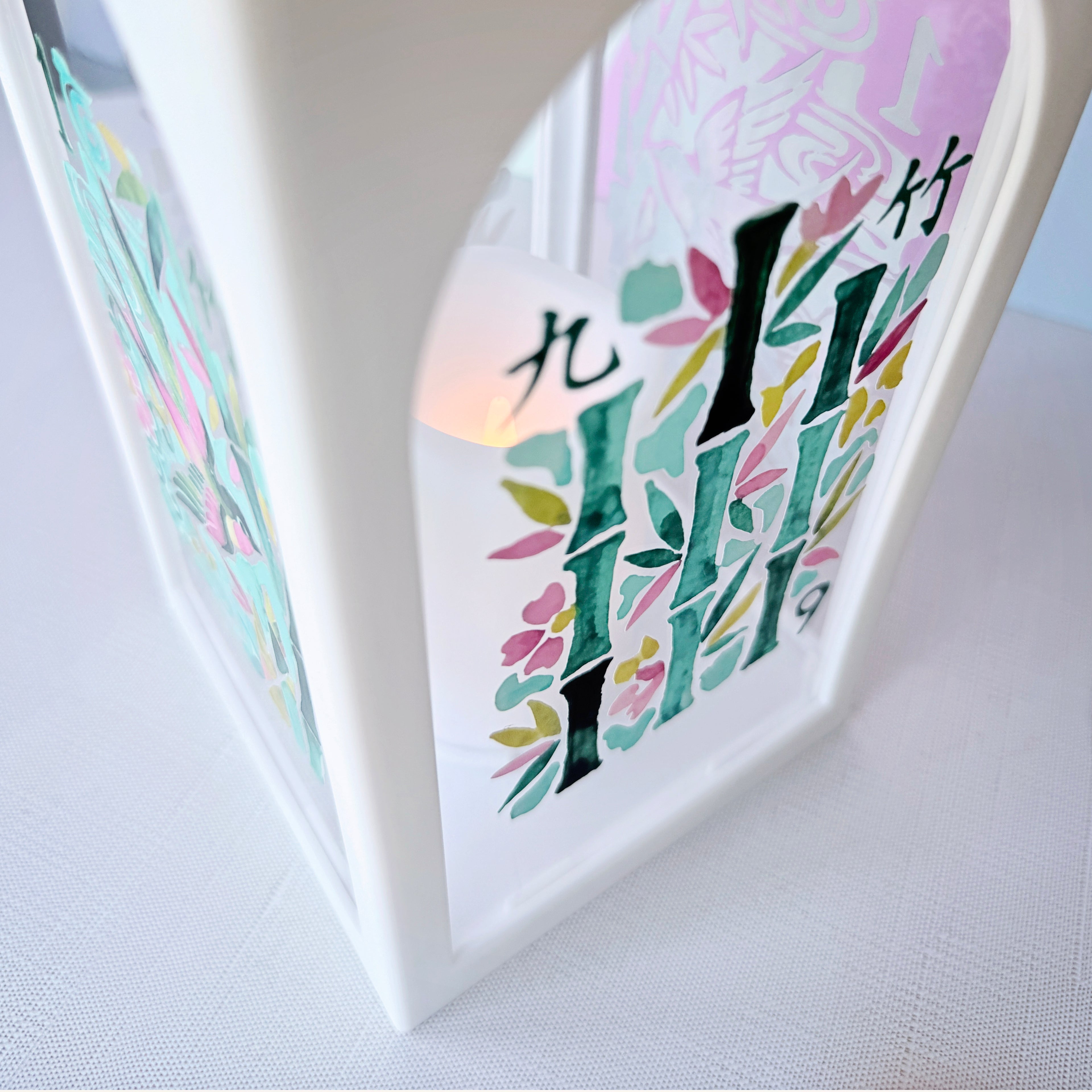 A close-up of a white decorative lantern with colorful printed bamboo and floral designs, lit from within, on a light fabric surface.