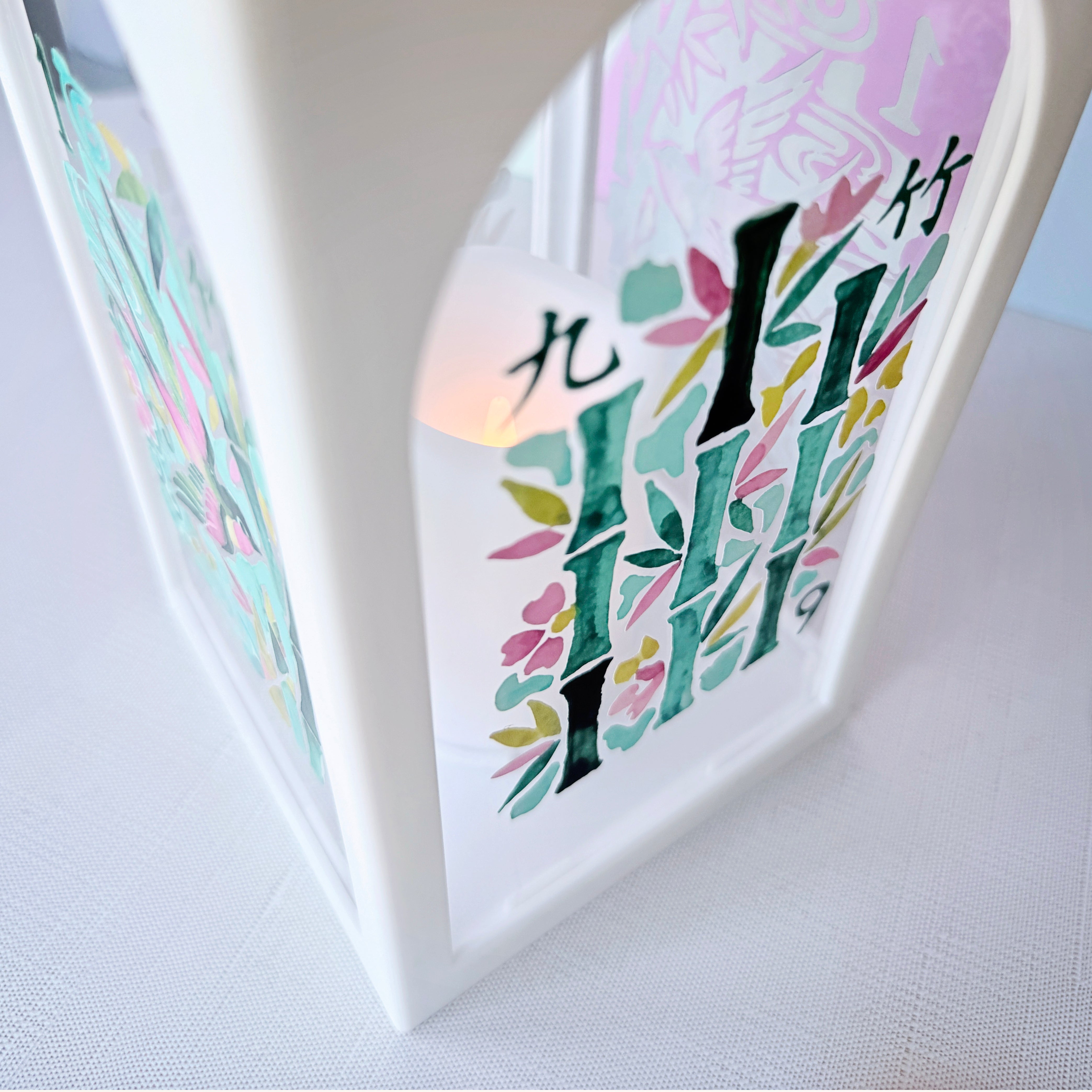 A close-up of a white decorative lantern with colorful printed bamboo and floral designs, lit from within, on a light fabric surface.