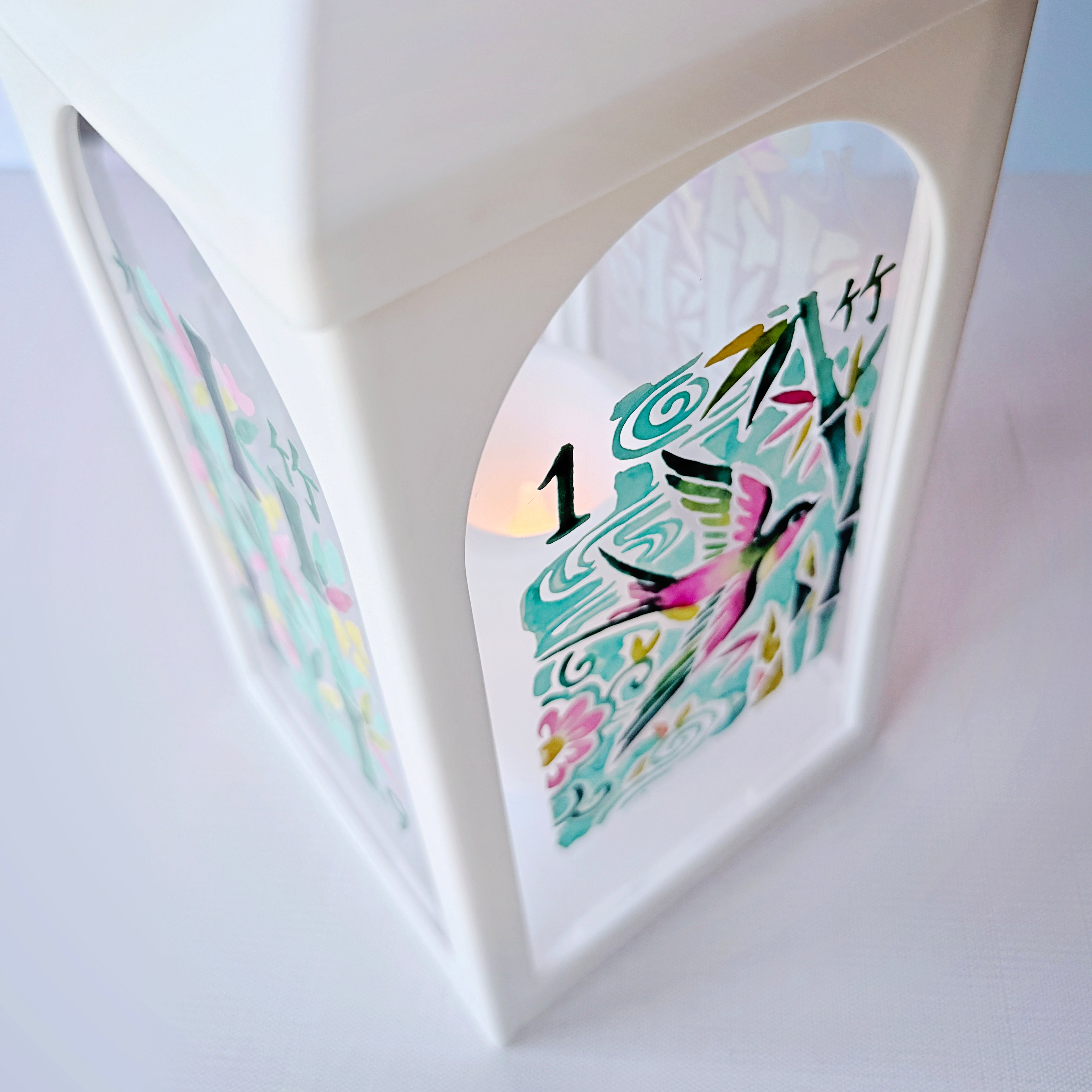 A white decorative lantern with an arched window displaying colorful Japanese-style artwork and the number 1, with a warm light glowing from within.
