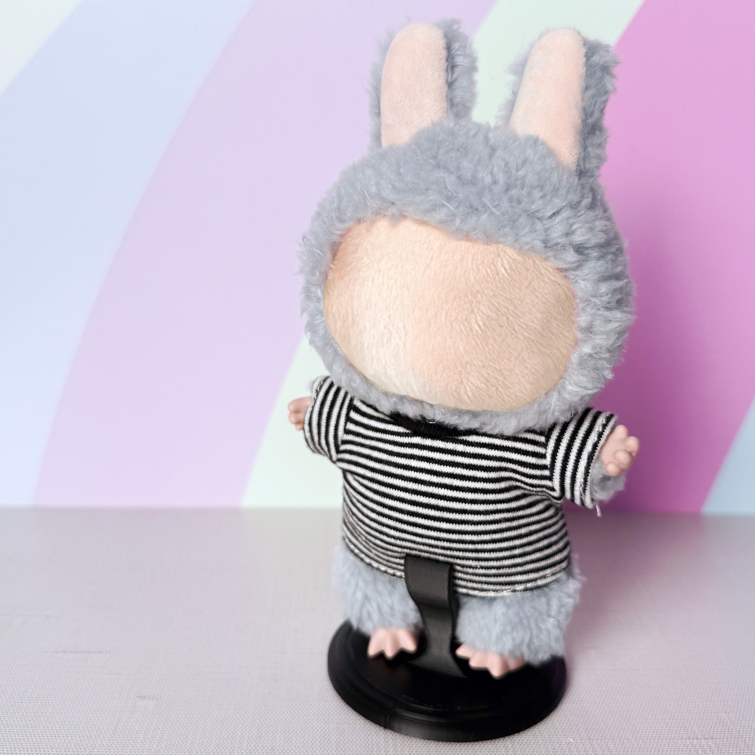 Plush toy with  black and white striped outfit on a black display stand against a pastel background.